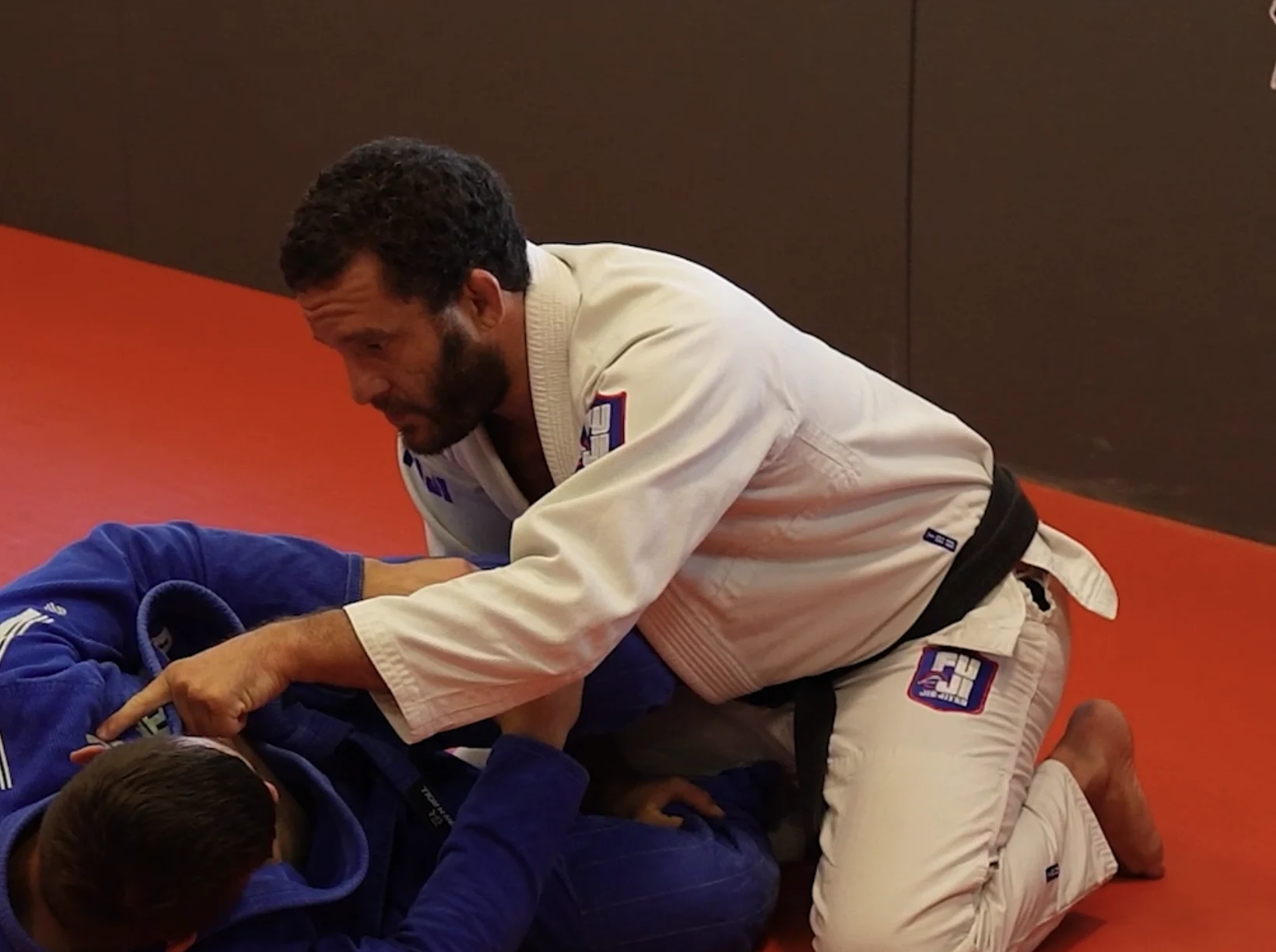 Two men practicing Brazilian jiu-jitsu, one in a white gi and the other in a blue gi, on a red mat indoors.