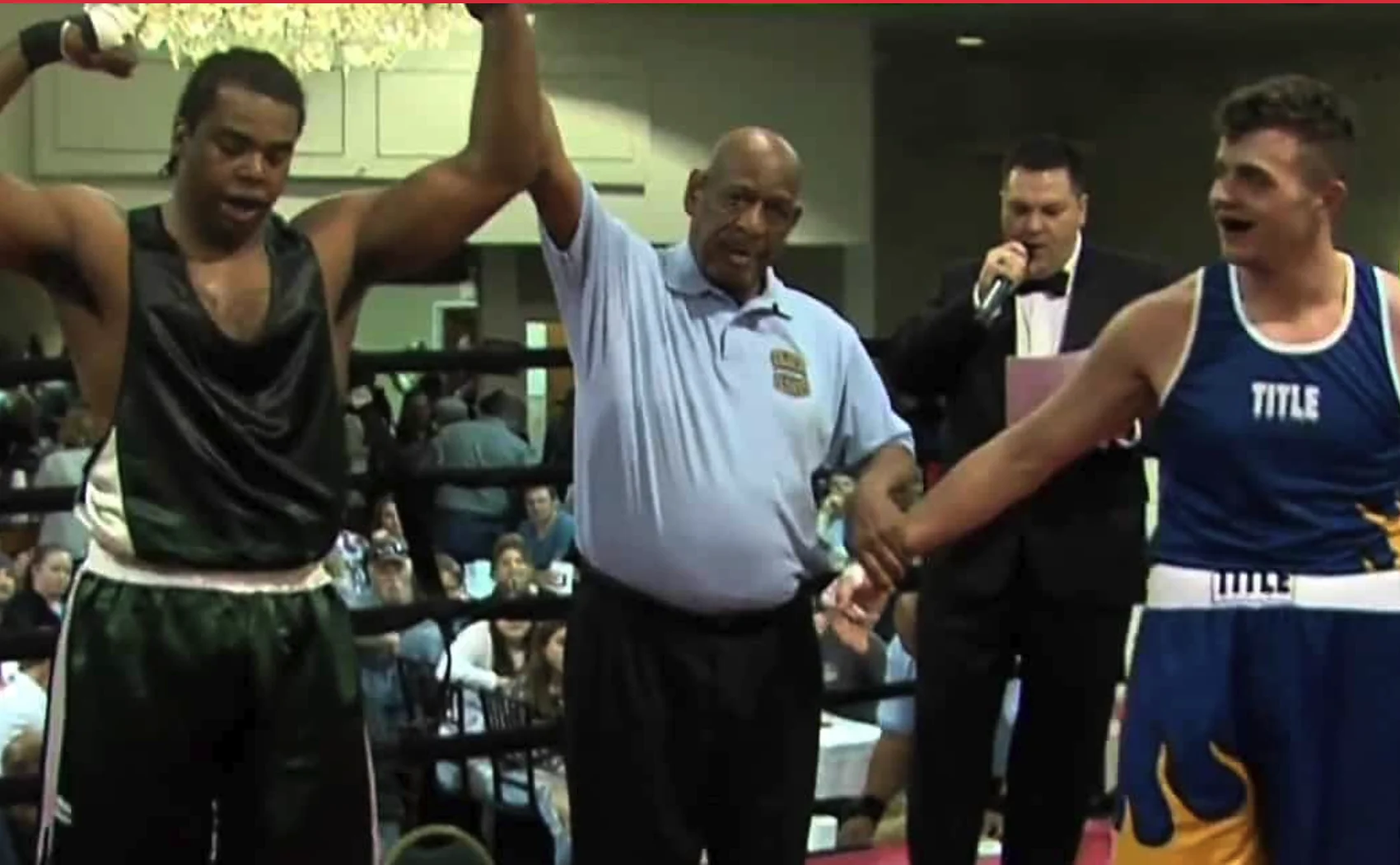 Boxer in black trunks raising his arm in victory as referee declares him the winner in a boxing ring, with the opponent in blue trunks looking surprised and a ring announcer in the background.